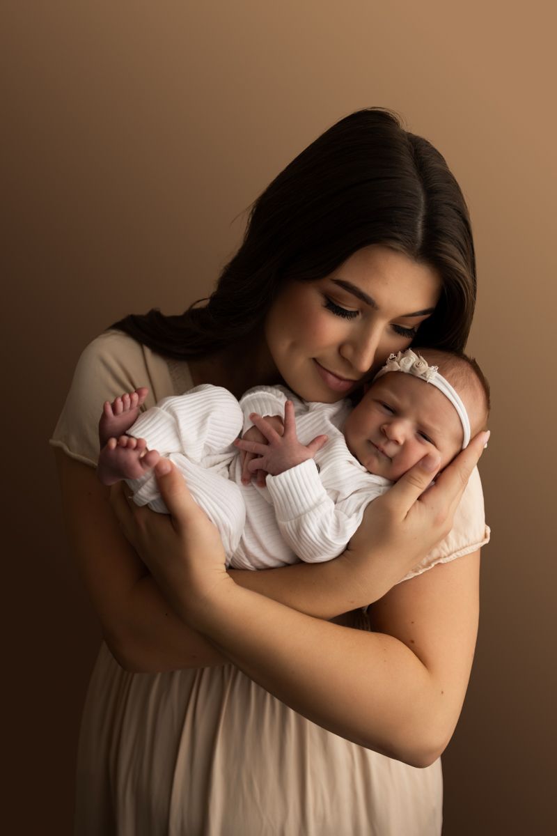 Newborn baby girl being held by her mom.  Baby is wearing a white onesie and mom is wearing tan dress.
