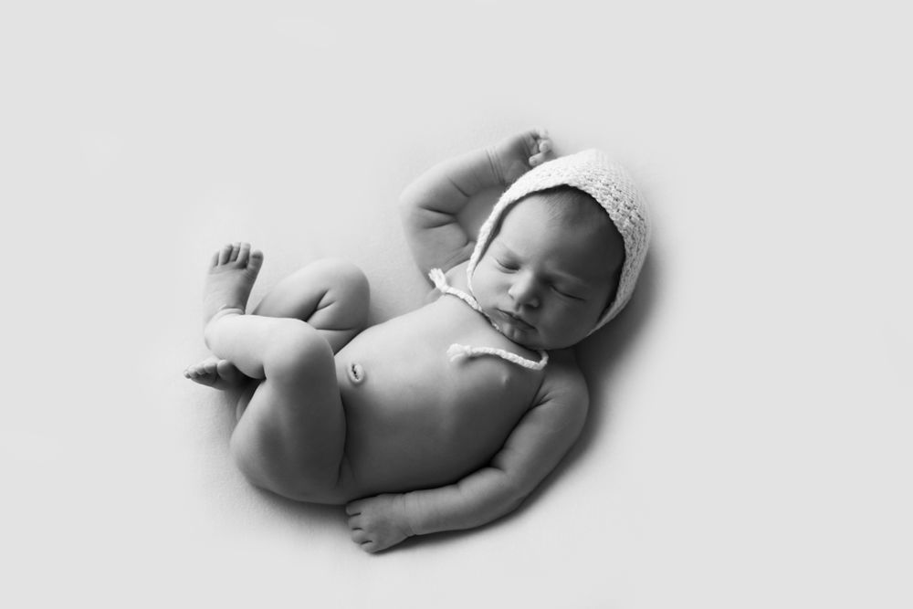 Black and white image of newborn baby girl in white bonnet on a white backdrop.
