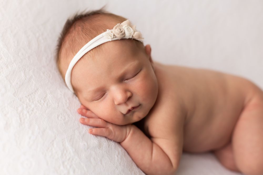 Newborn baby girl with her hand under the cheek and white headband.