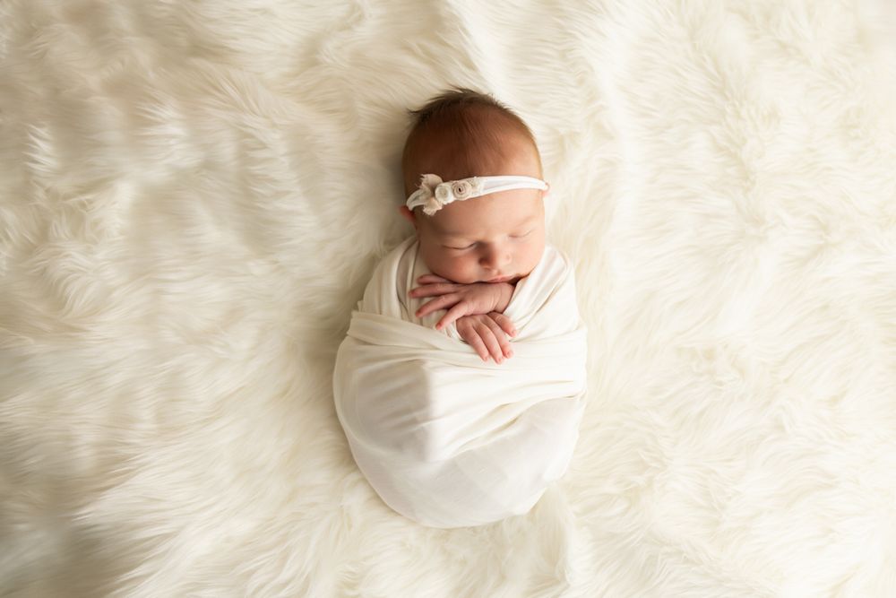 Newborn baby girl wrapped in a white wrap and headband on fur blanket.