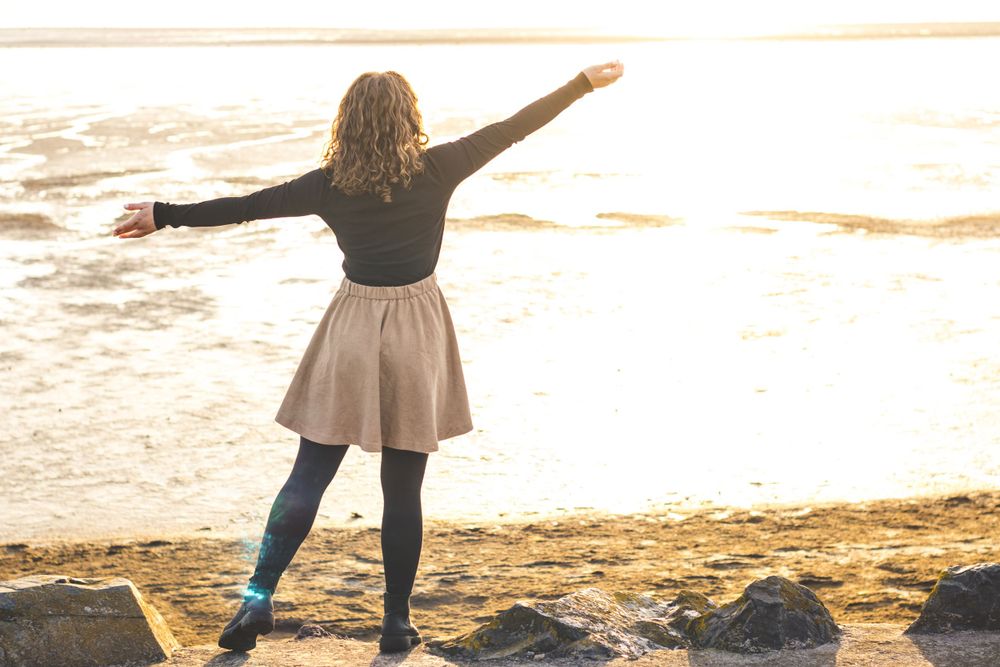 A silhouette figure stands with arms outstretched on rocky beach at sunrise