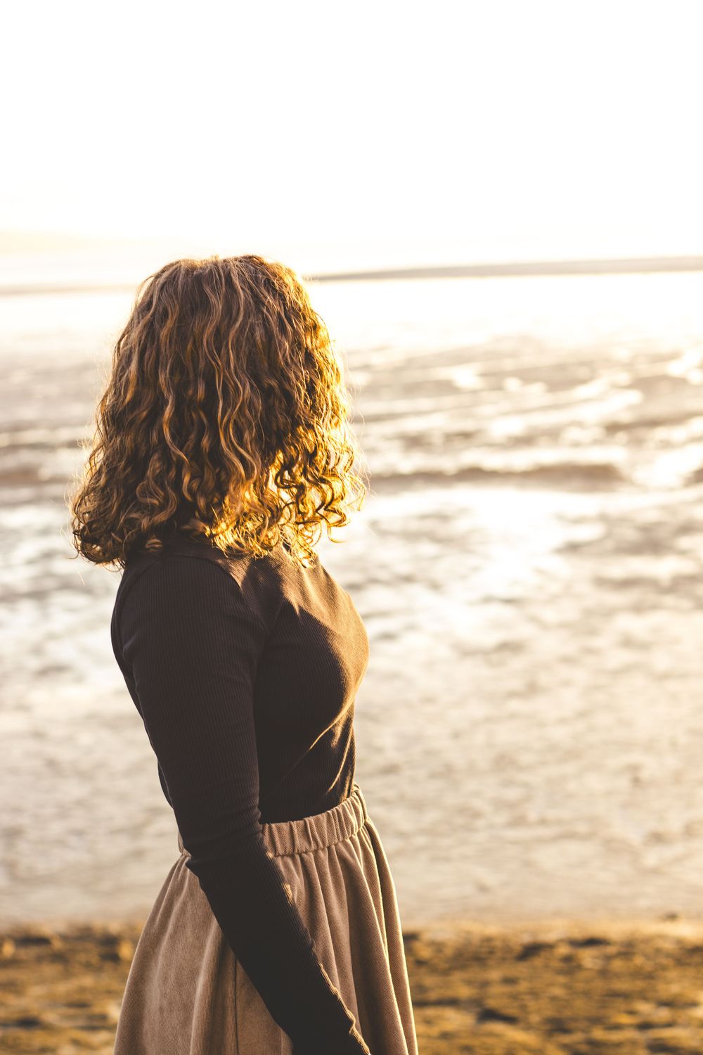 Person with curly hair gazes at ocean waves during golden hour sunrise.