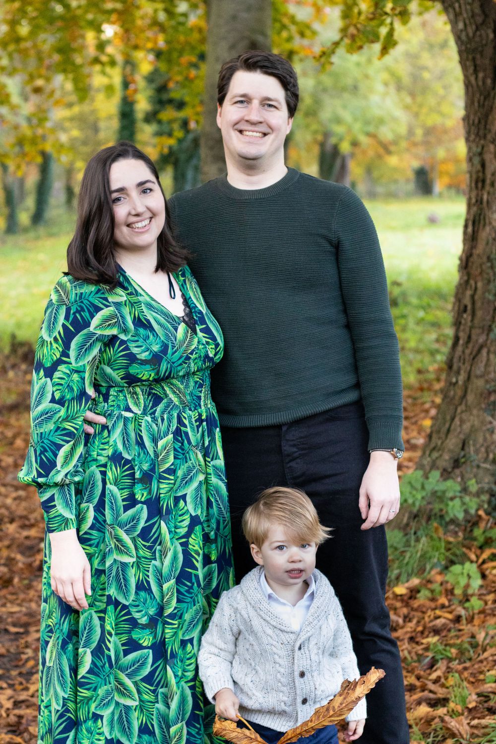 Family of three poses together in autumn park with fallen leaves.