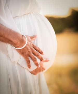 Pregnant woman in white dress gently cradling baby bump with hands wearing white bracelet outdoors.