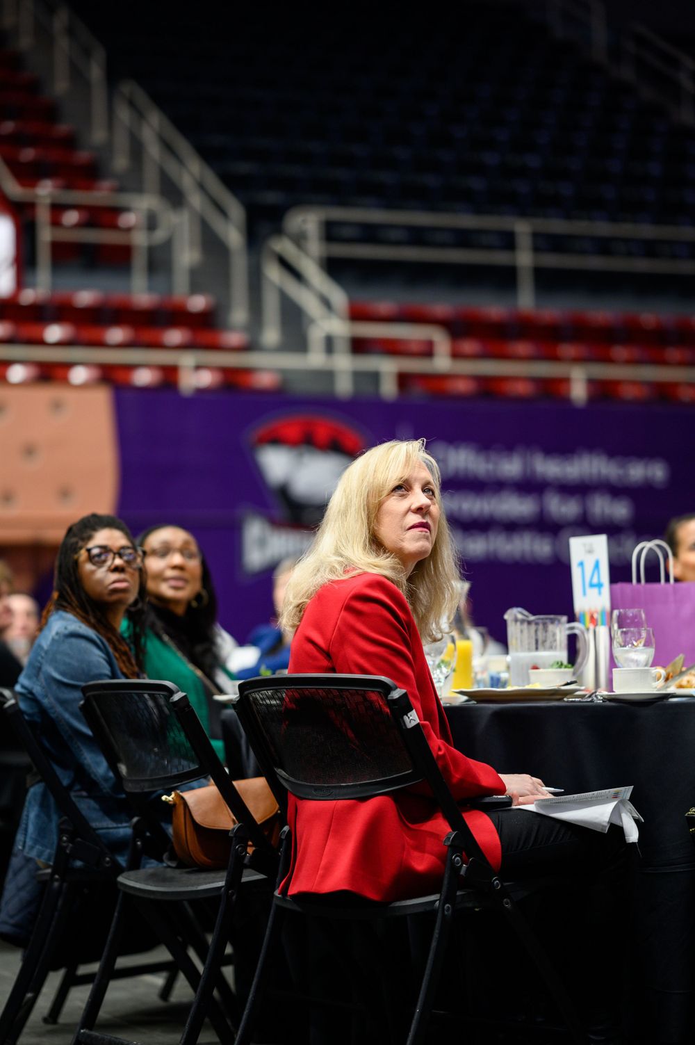 Attendees sitting at round tables during an indoor event with stadium seating in background.