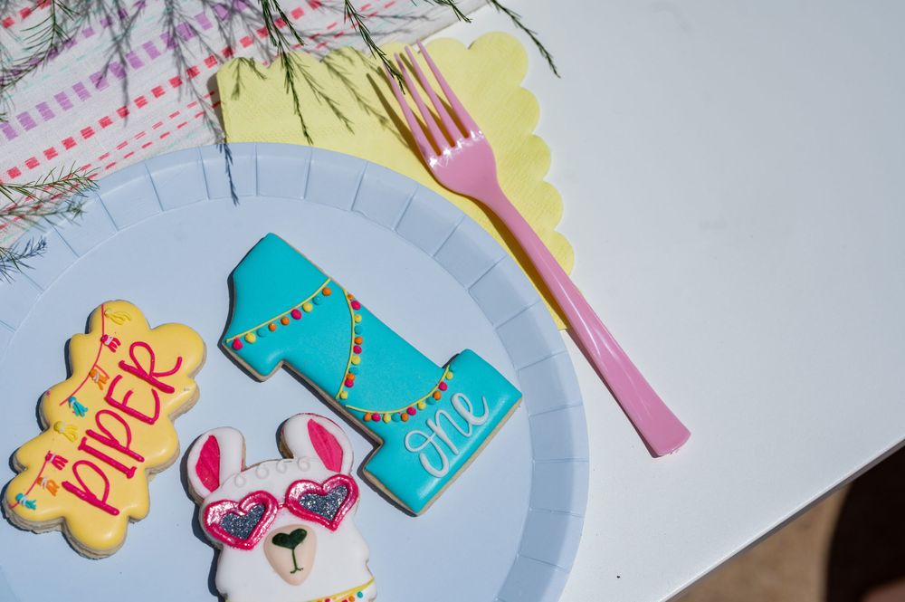 Decorative cookies and party supplies including a pink fork and turquoise number one on a plate.