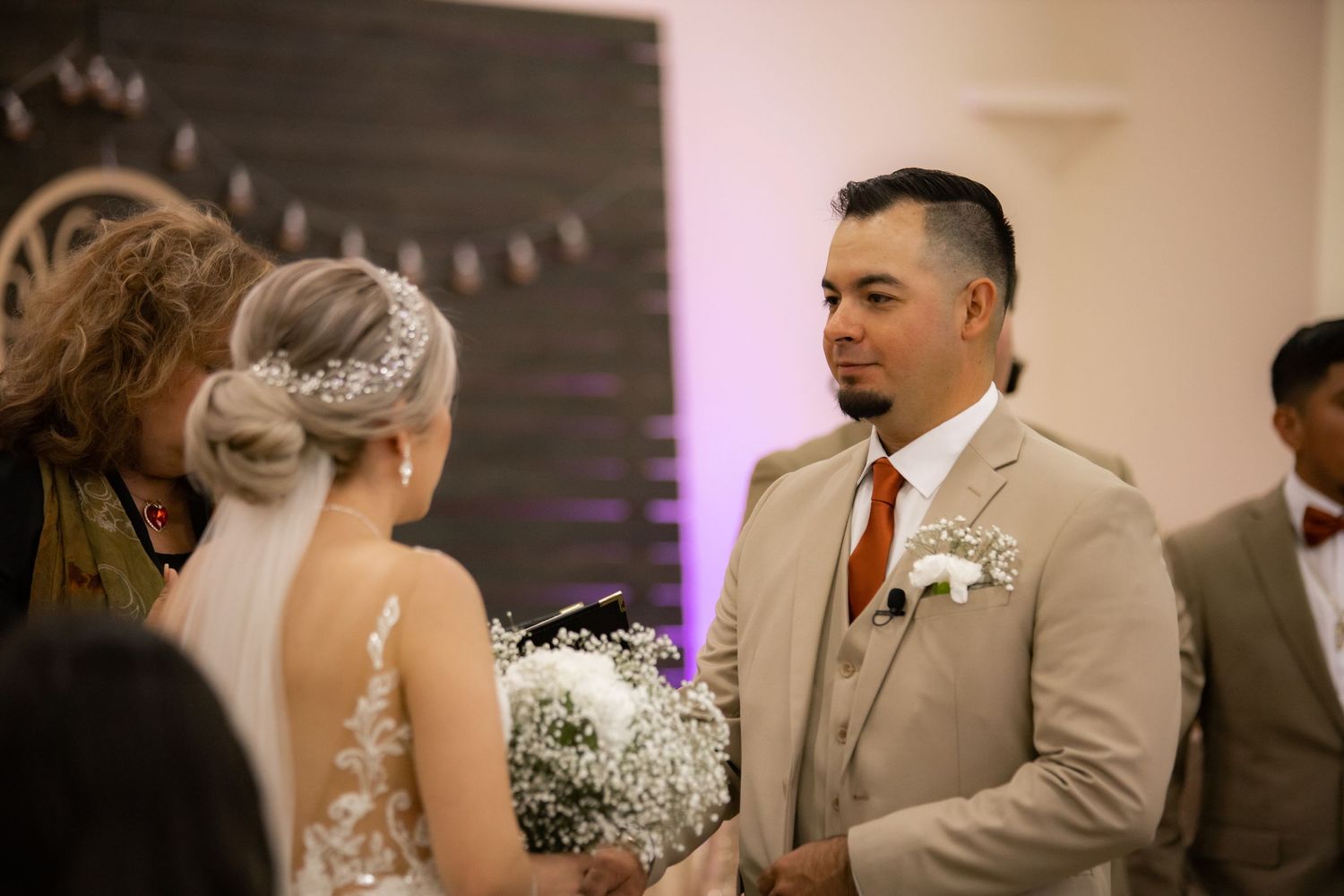 Groom in a beige suit exchanging vows with bride holding a white bouquet during indoor wedding ceremony.