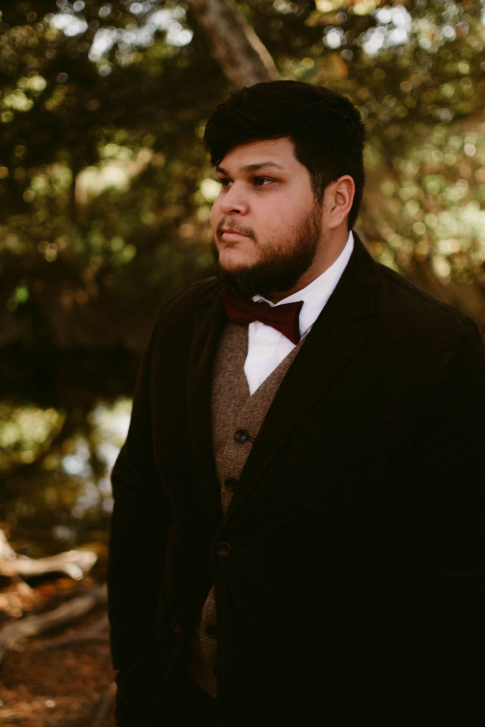 Classic formal portrait in black tuxedo and brown vest against forest backdrop.