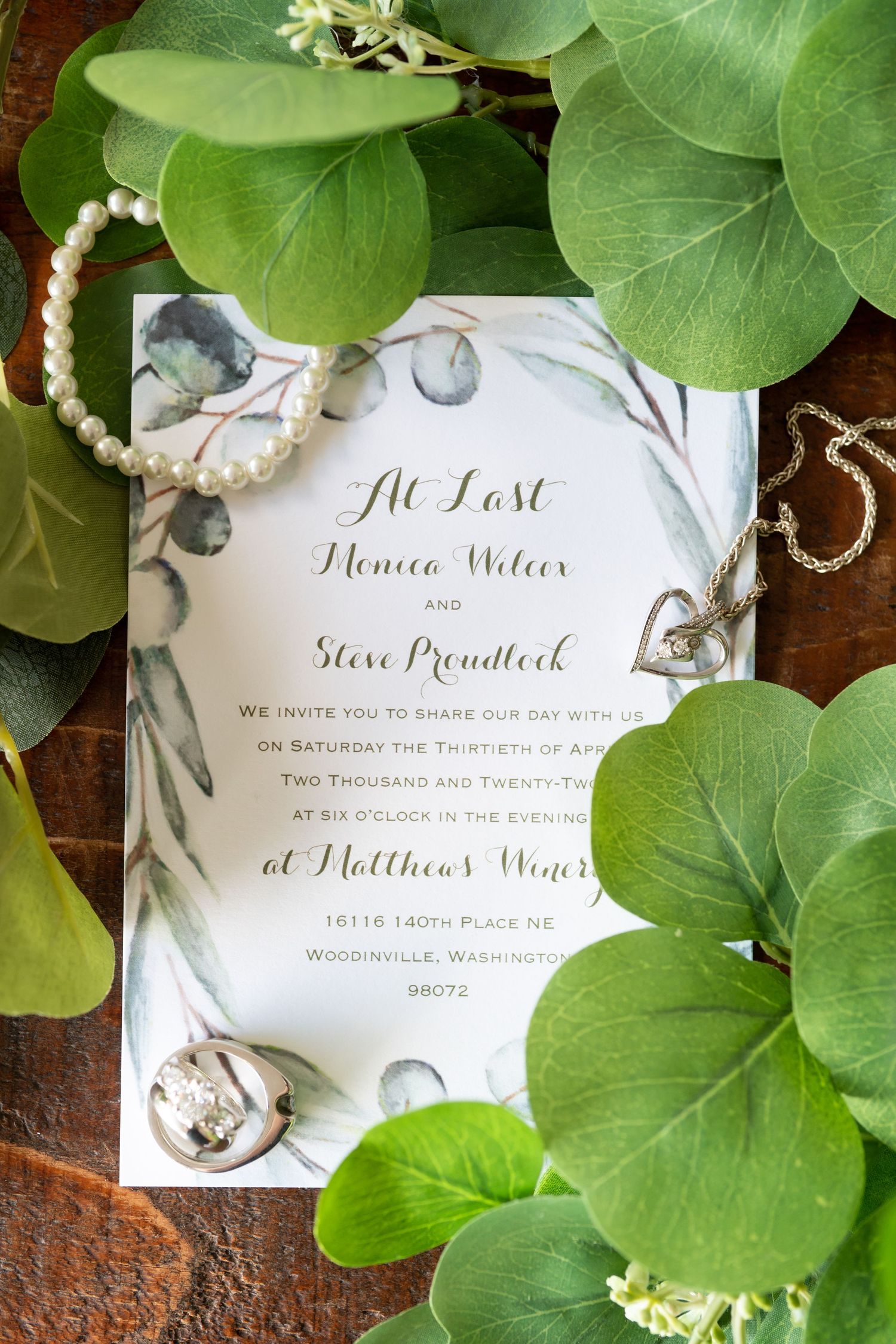 An elegant wedding invitation surrounded by green leaves and pearl accessories.