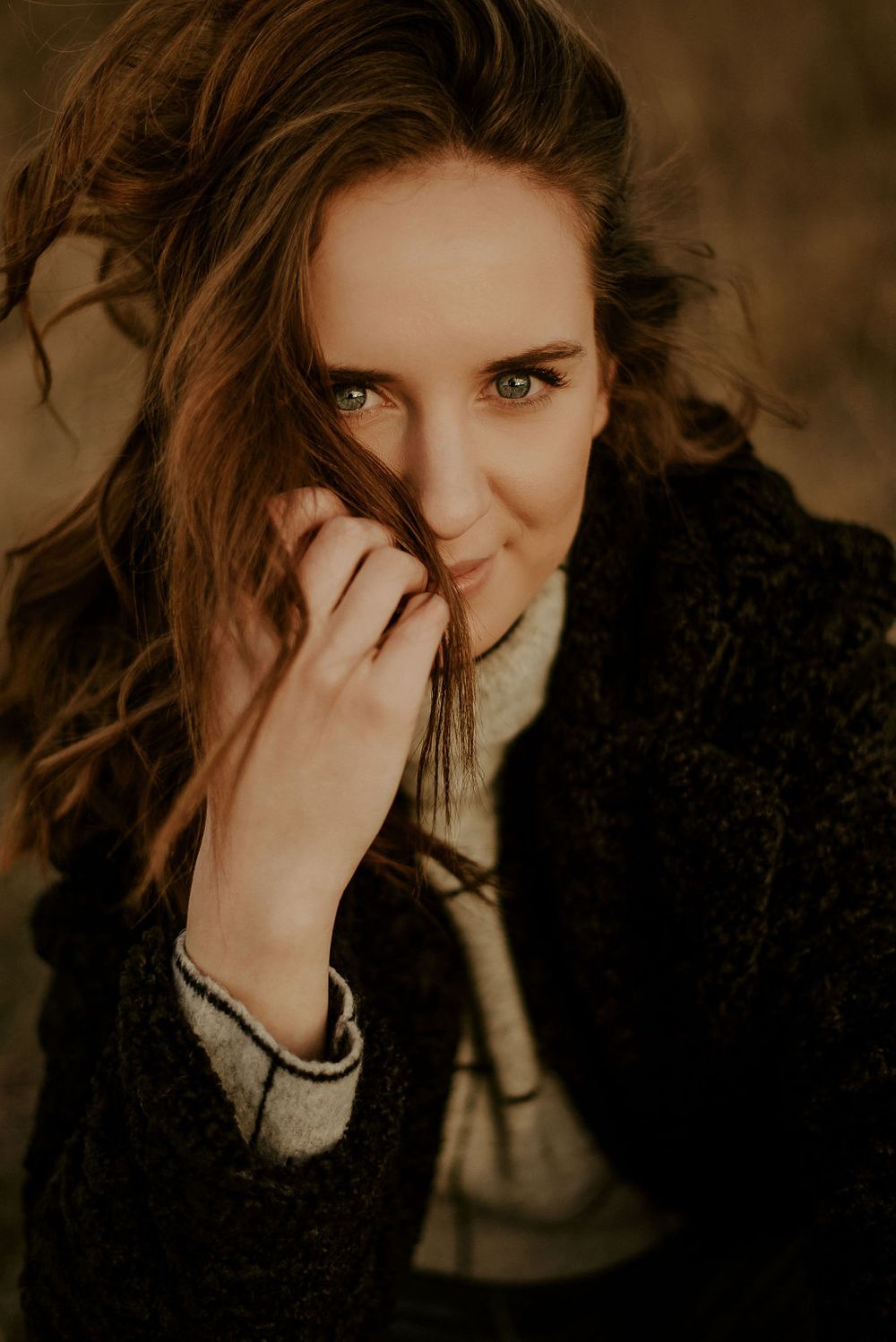A portrait of someone in a black coat with windswept hair and playful expression in warm lighting.