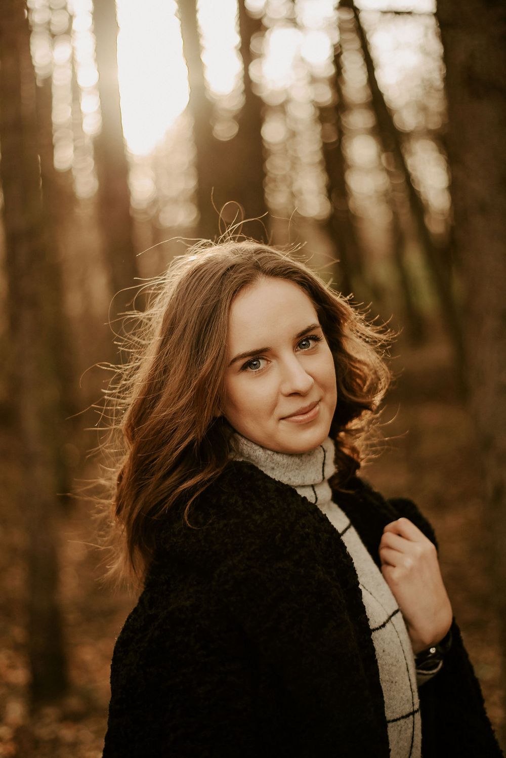Artistic outdoor portrait in warm autumn light with windswept hair and black coat against blurred forest background.