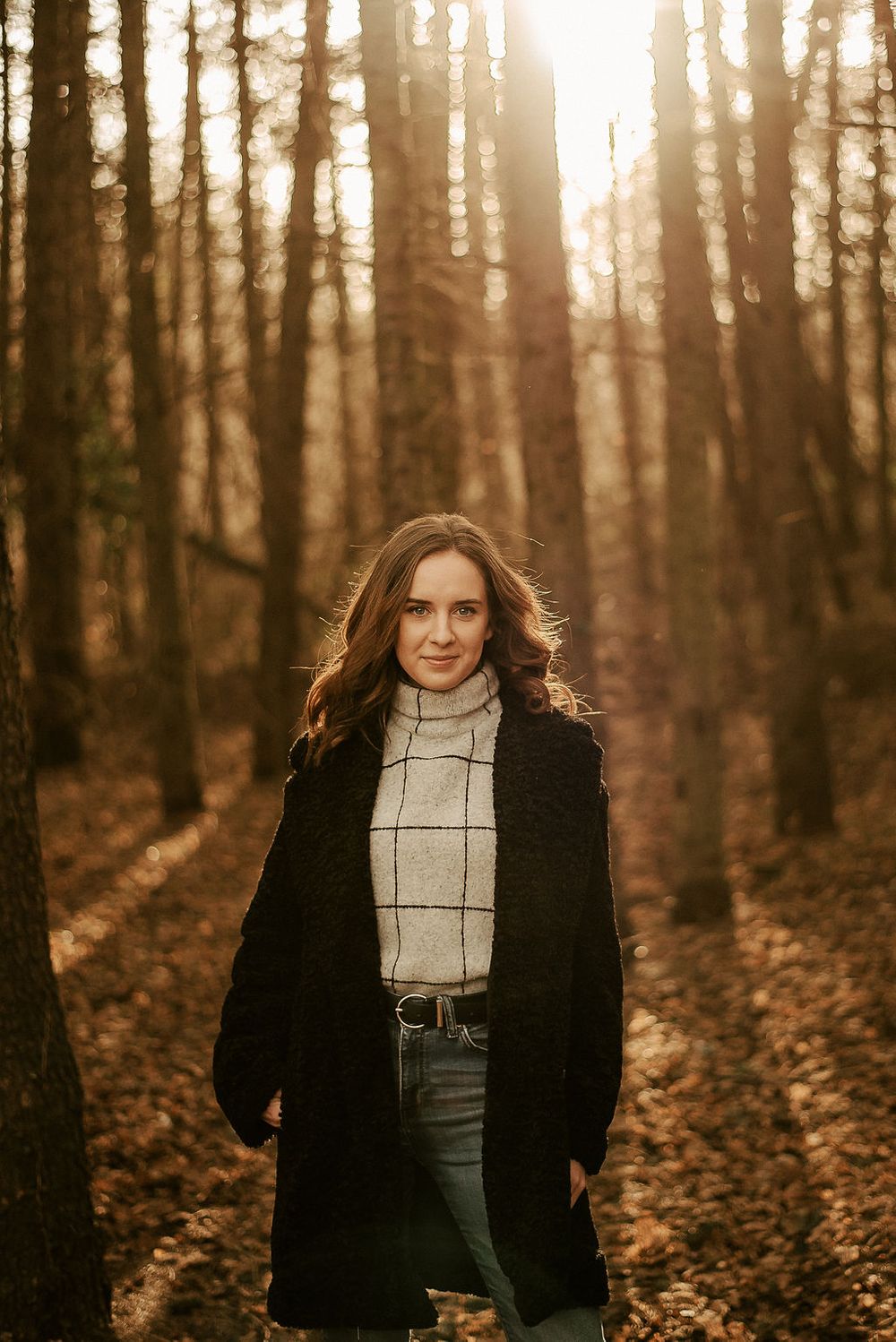 Sunlit forest portrait with black coat and plaid turtleneck against golden trees.