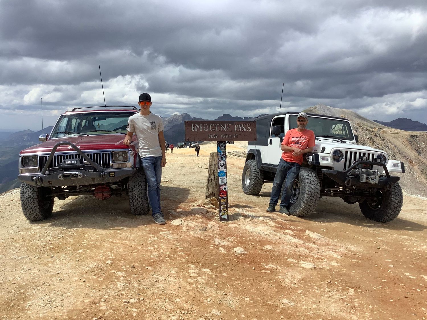 Two men posing with off-road Jeeps at a mountain pass sign under cloudy skies.