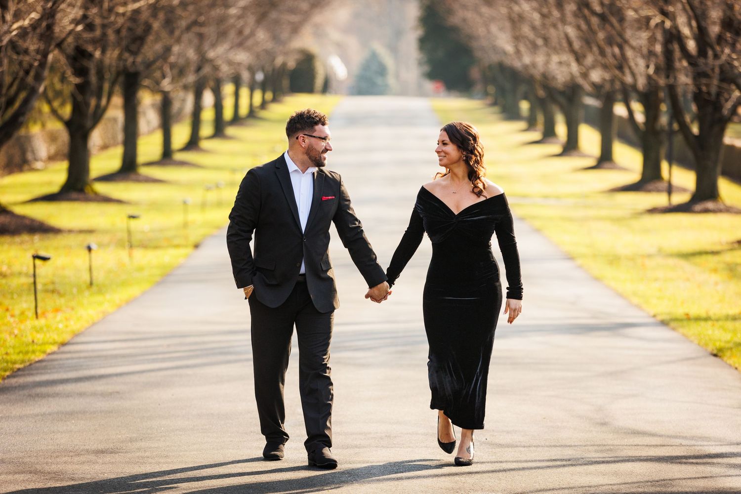 Stylish couple holding hands walking down a scenic tree-lined avenue in formal attire.