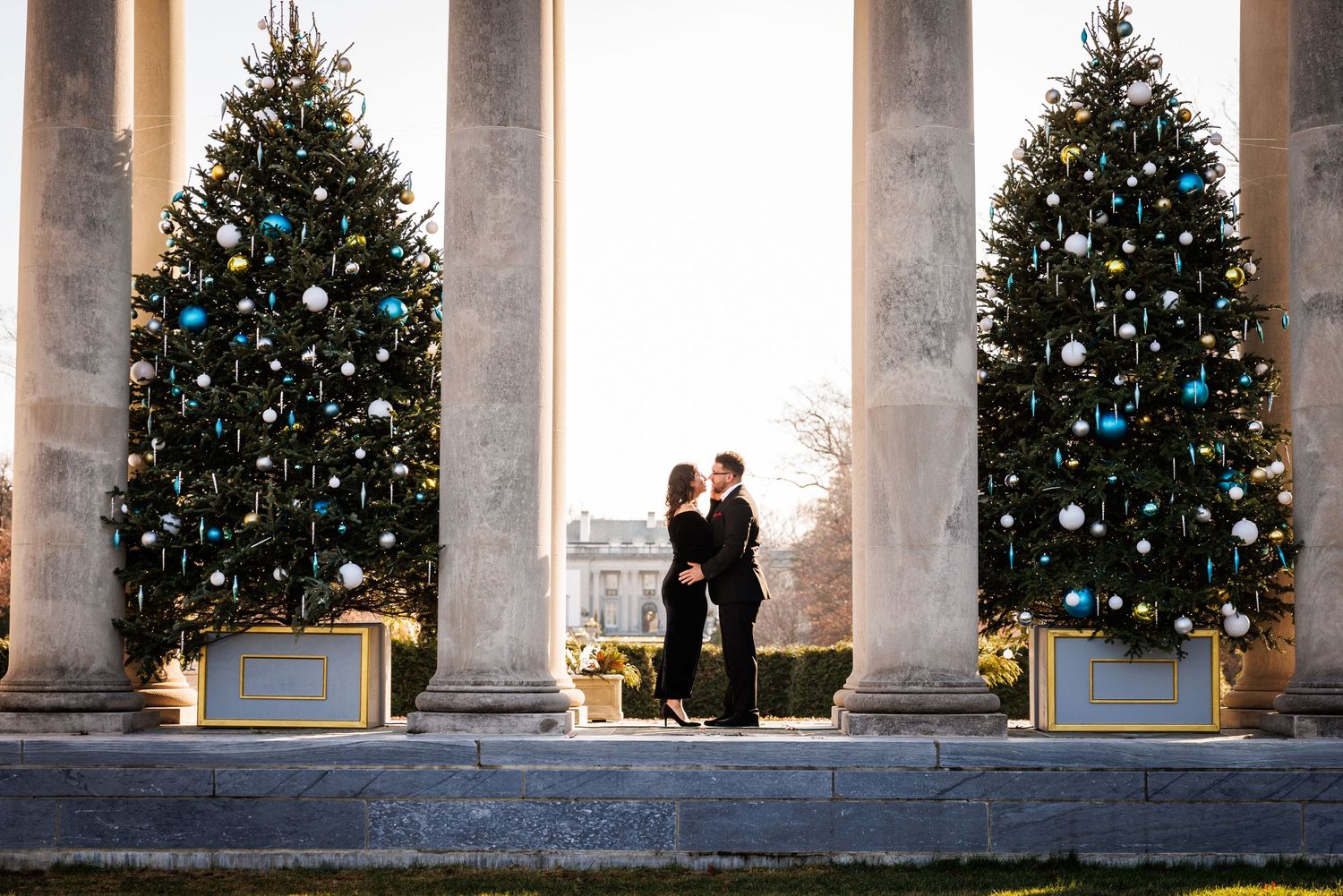 Couple kissing between two decorated Christmas trees framed by grand columns at dusk.