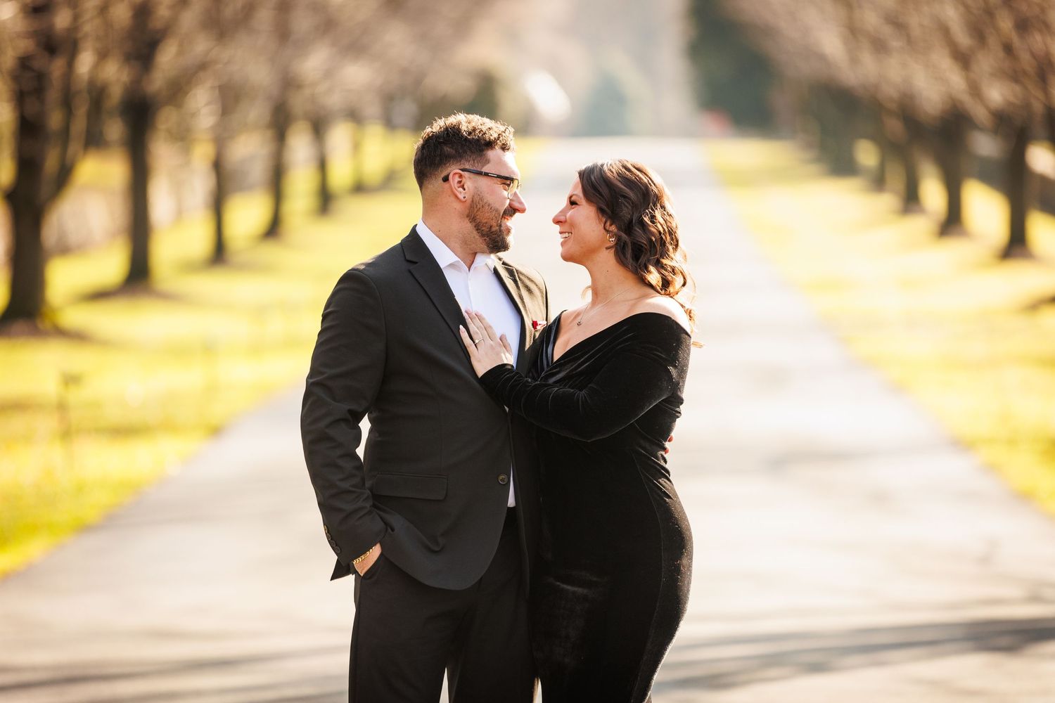Elegant couple in formal black attire gazing at each other on a tree-lined path.
