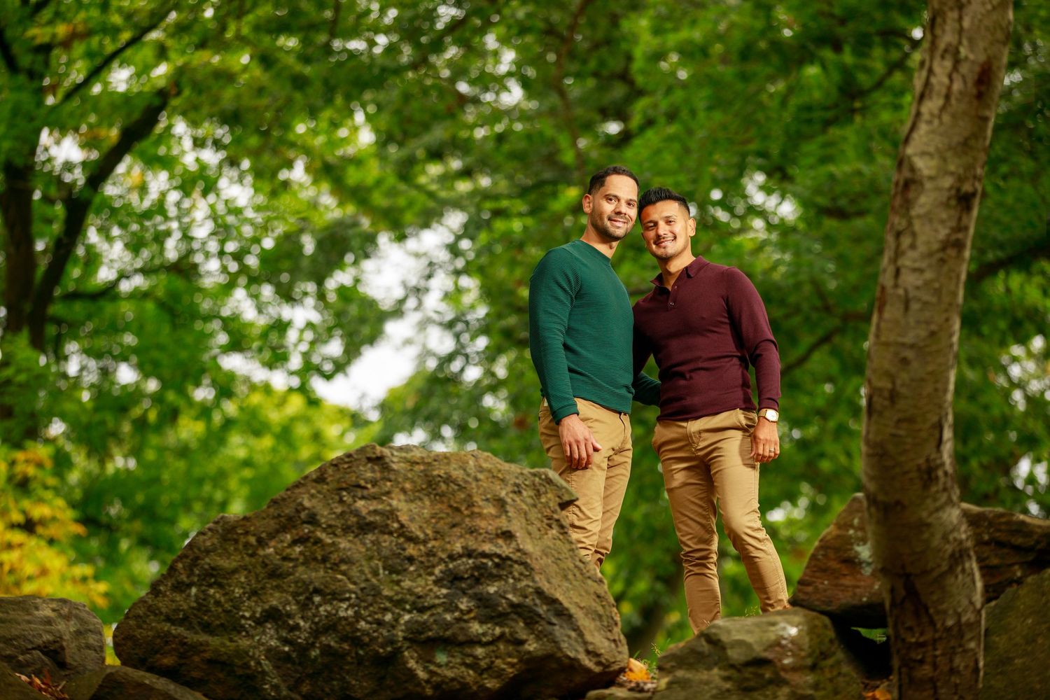 Two men in coordinating sweaters smile together while standing on large rocks in a lush green park.