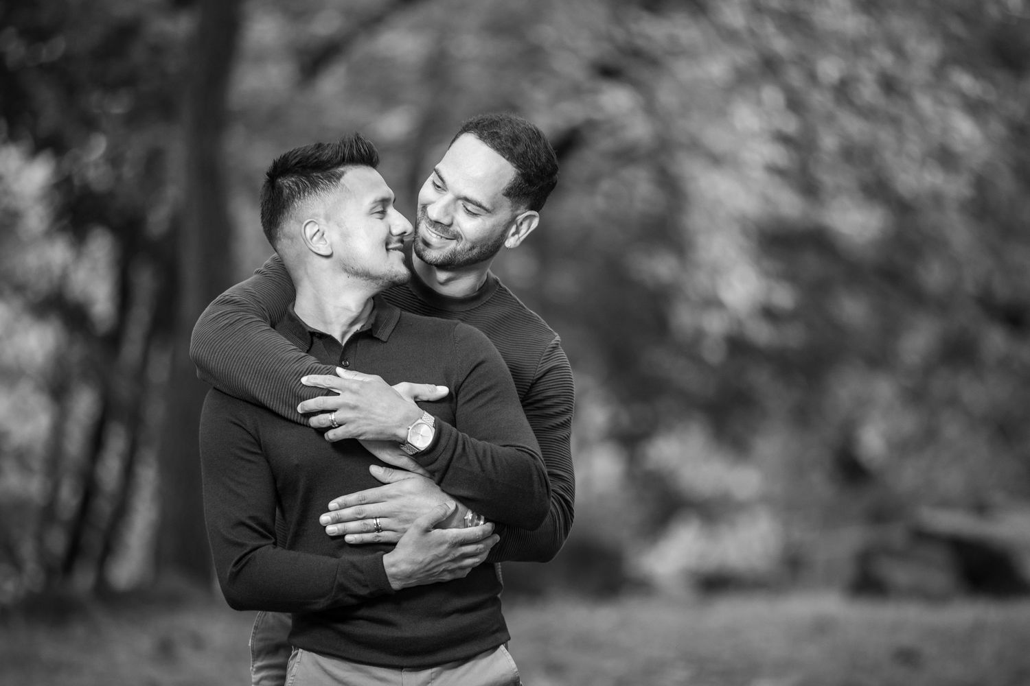 Two men share a loving embrace in a black and white outdoor portrait among autumn trees.