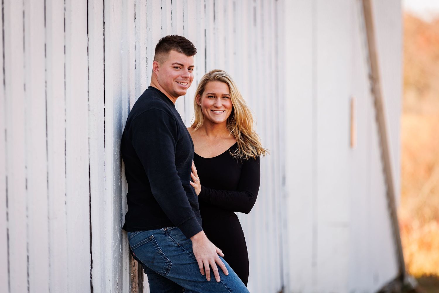 Smiling couple leaning against white barn wall in warm autumn sunlight.