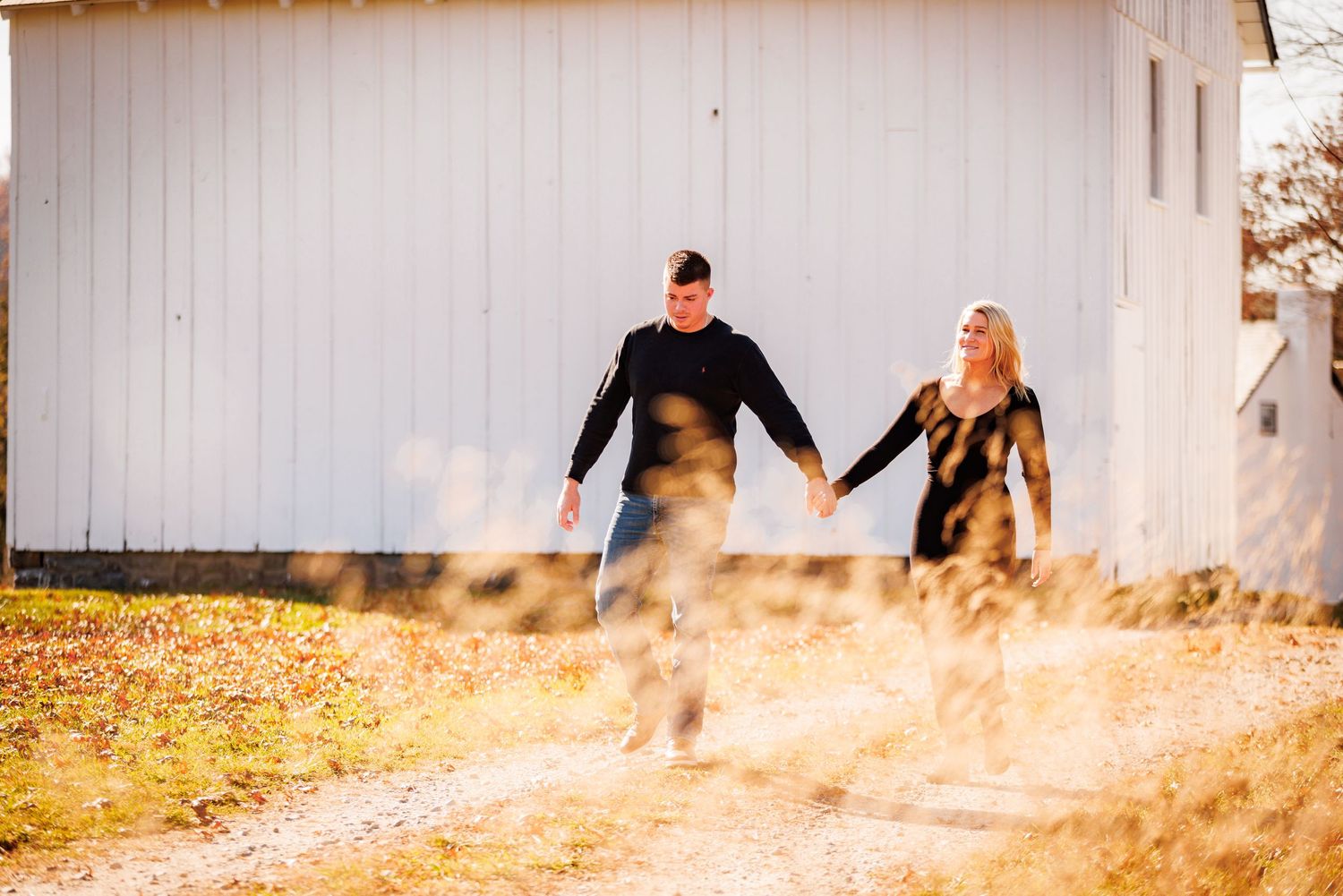 Happy couple holding hands walking along dirt path beside white barn in fall.