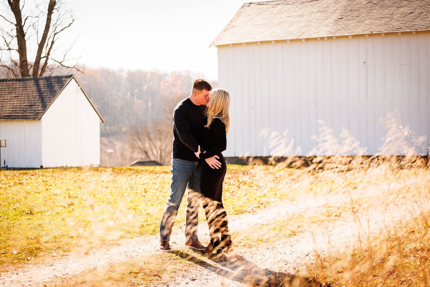 Couple sharing a romantic kiss on a sunny path beside a white barn in fall.