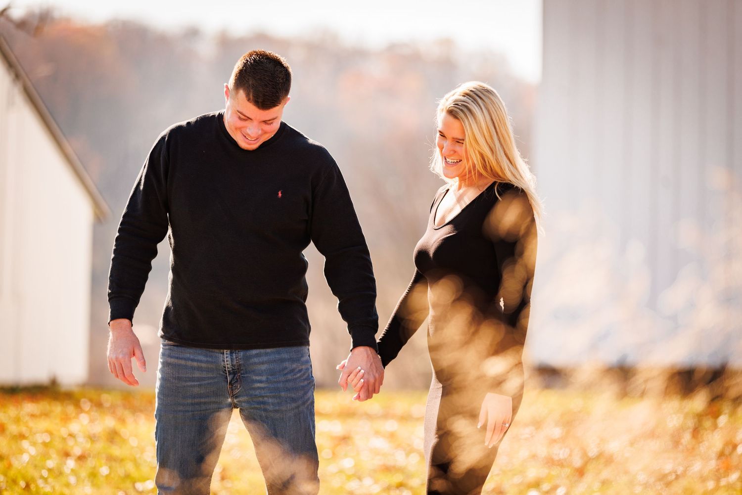Couple holding hands and walking through golden autumn field beside white barn.