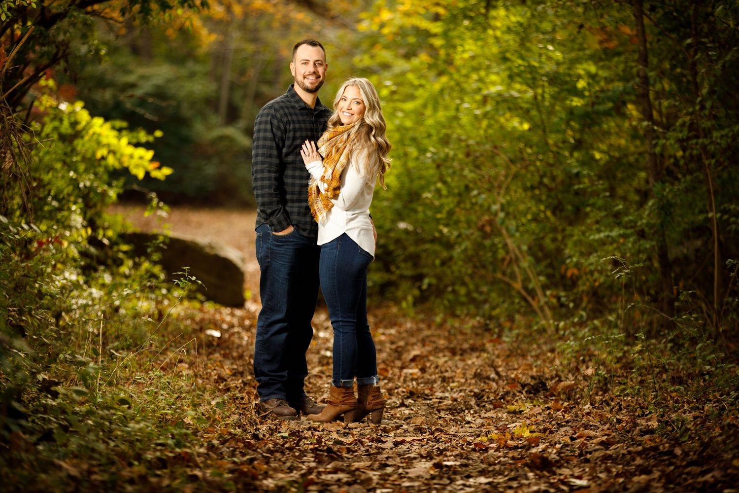 Smiling couple posing on a leaf-covered path surrounded by vibrant autumn foliage.