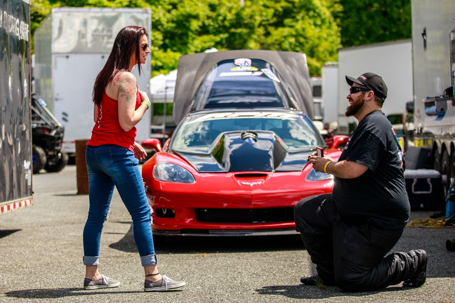 Man kneeling to propose to woman in front of a red Corvette at an outdoor car event.