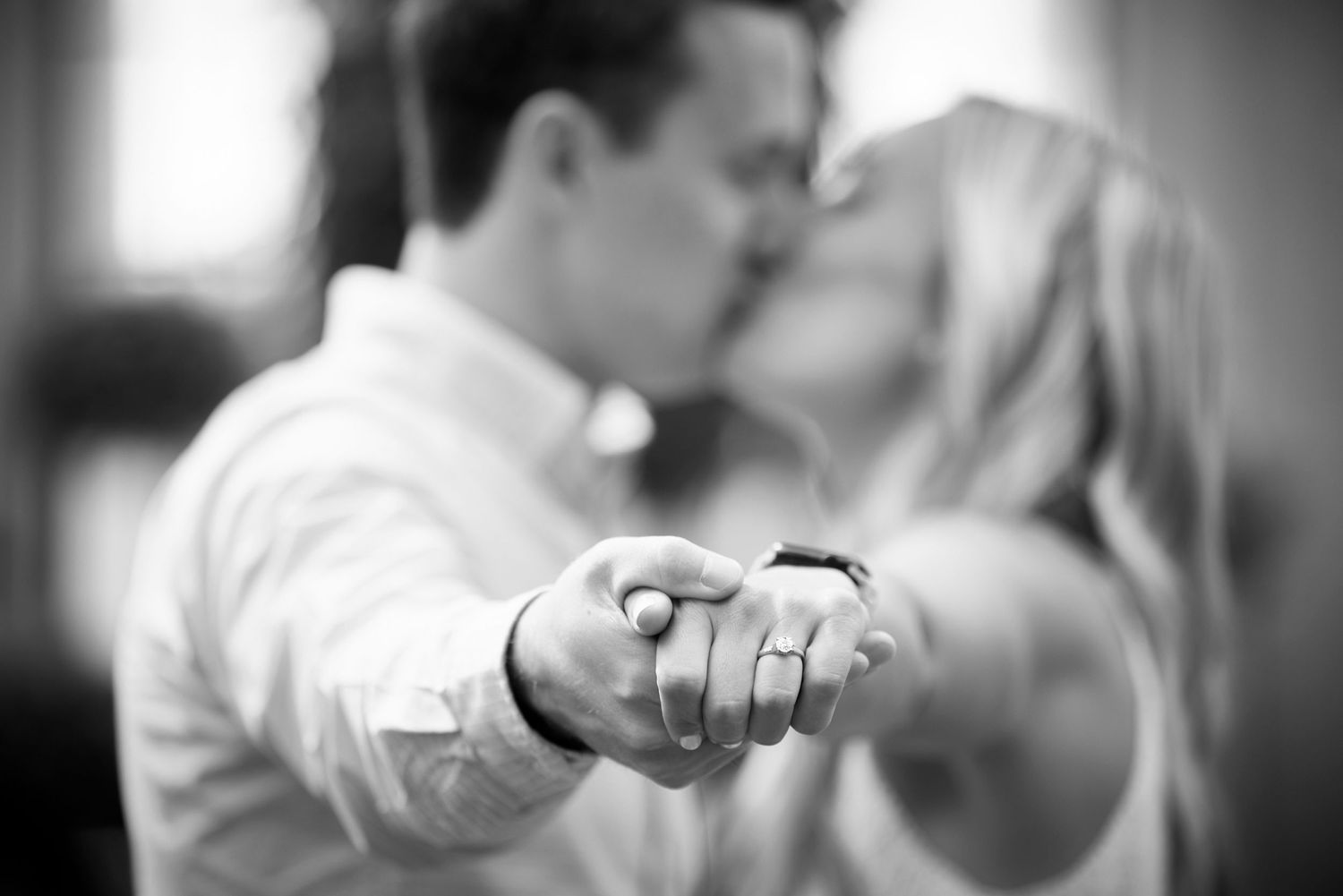 Black and white close-up of kissing couple holding hands toward camera showing rings.