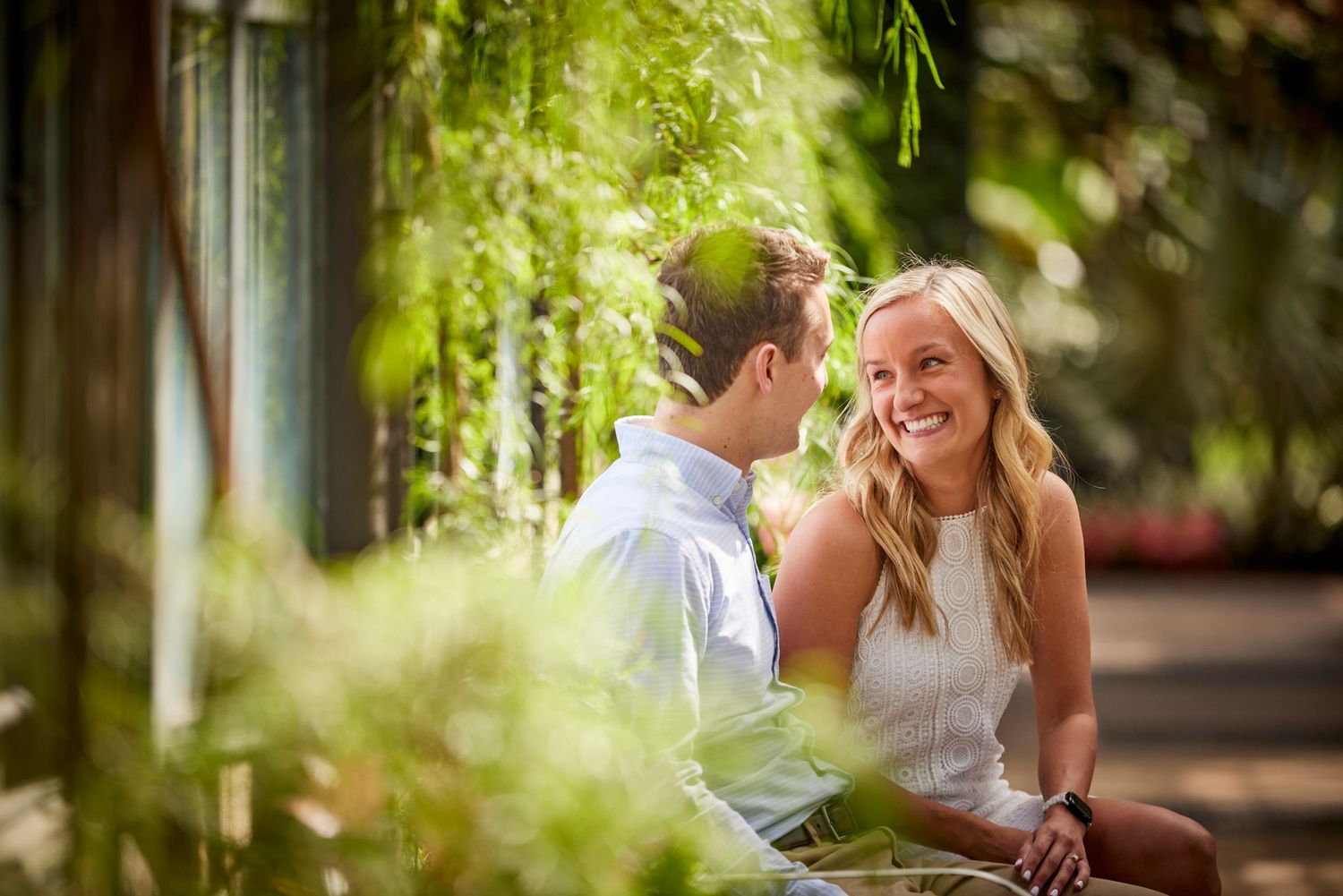 Smiling blonde woman sitting with partner amid lush green garden foliage outdoors.