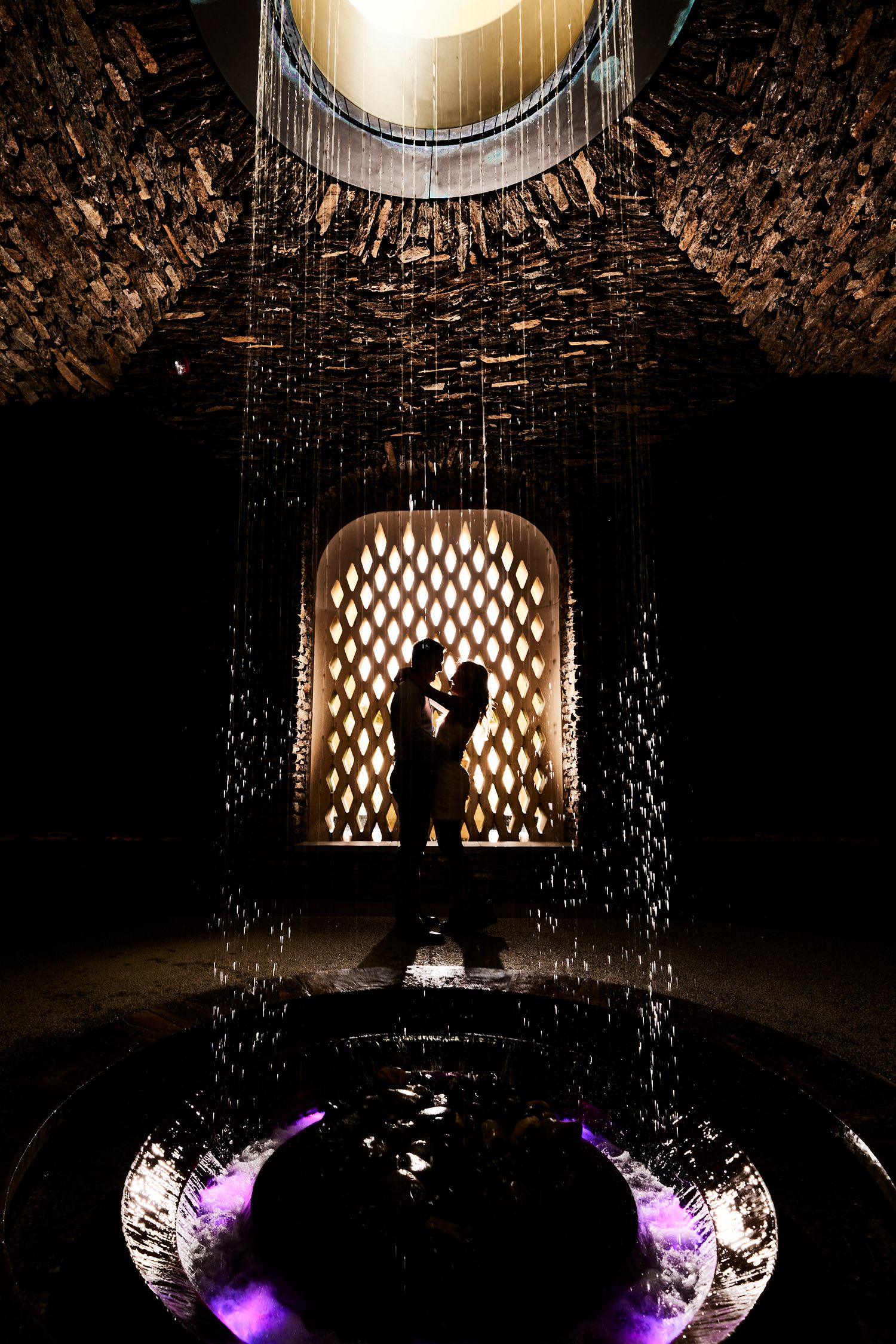 Romantic silhouette of couple embracing in dramatic lit archway with cascading water curtain.