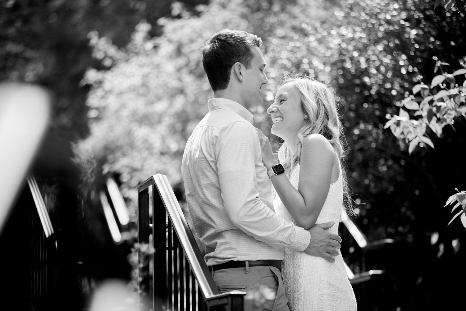 Black and white photo of smiling couple embracing outdoors near garden staircase.