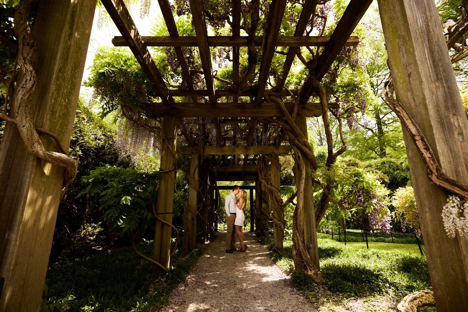 Couple walking hand-in-hand through a lush garden pergola covered in climbing vines.