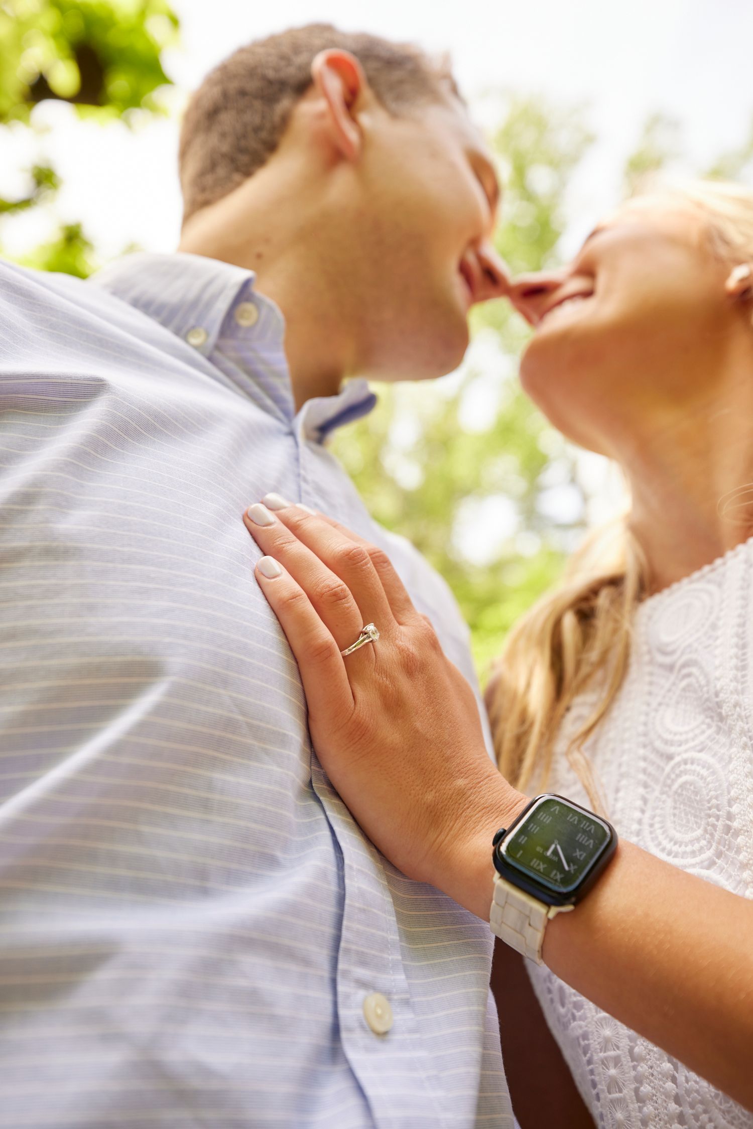 Couple sharing a tender kiss outdoors, woman's hand on man's chest displaying engagement ring.