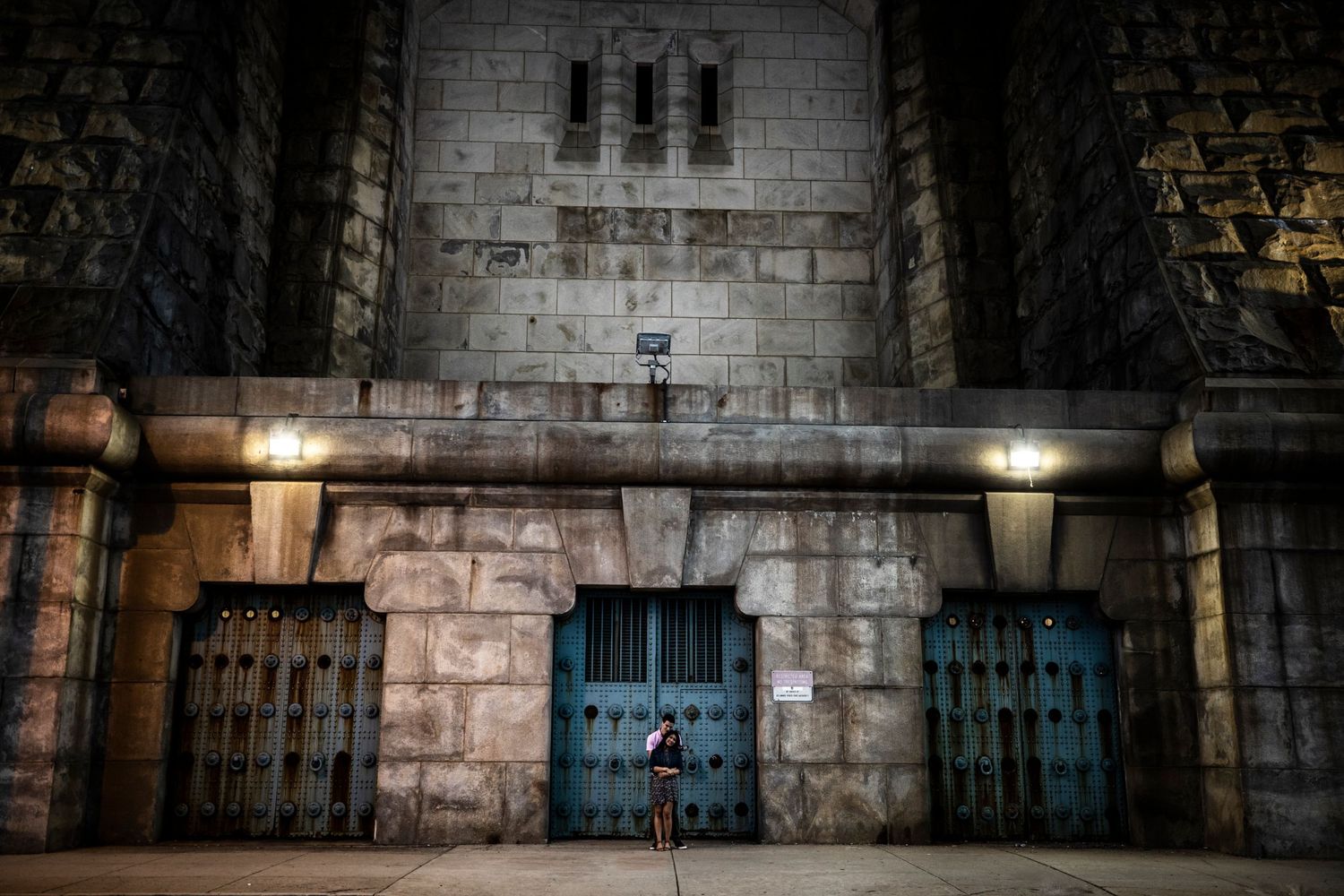 A lone figure stands before a massive stone building with ornate blue doors at night.