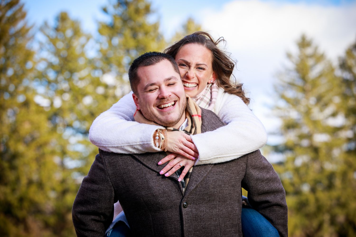A smiling couple shares a joyful piggyback ride outdoors with evergreen trees in the background.