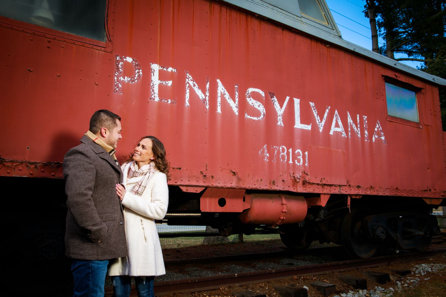 An engaged couple shares a romantic moment beside a vintage red Pennsylvania railroad car.