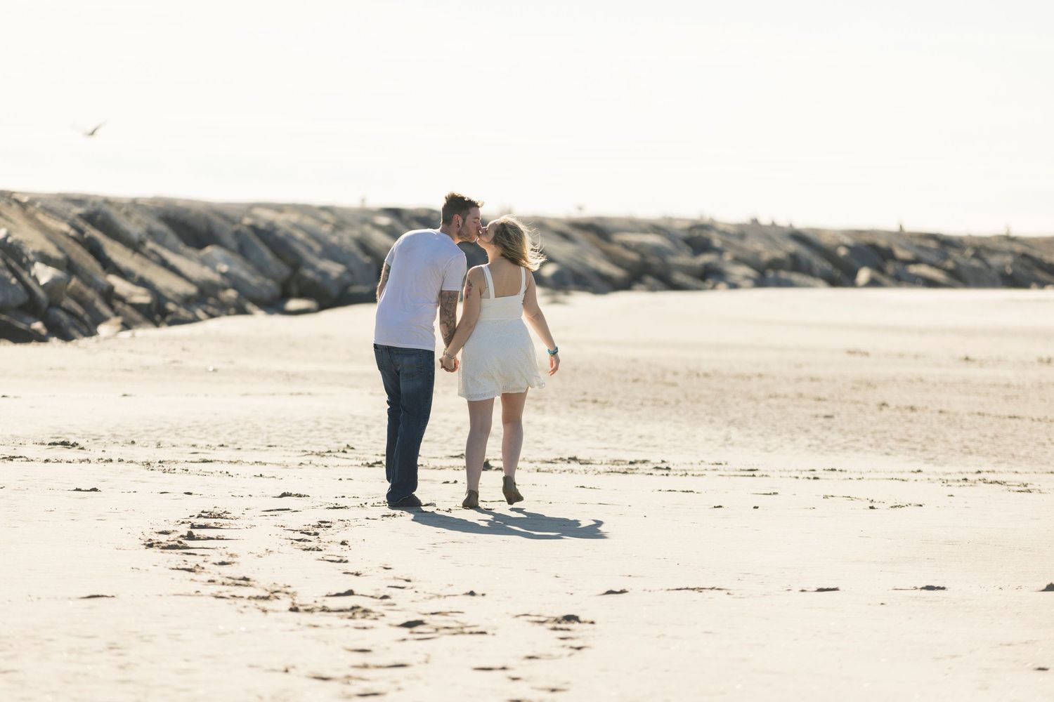 Couple walking hand in hand on a sandy beach, sharing a kiss near a rocky jetty.