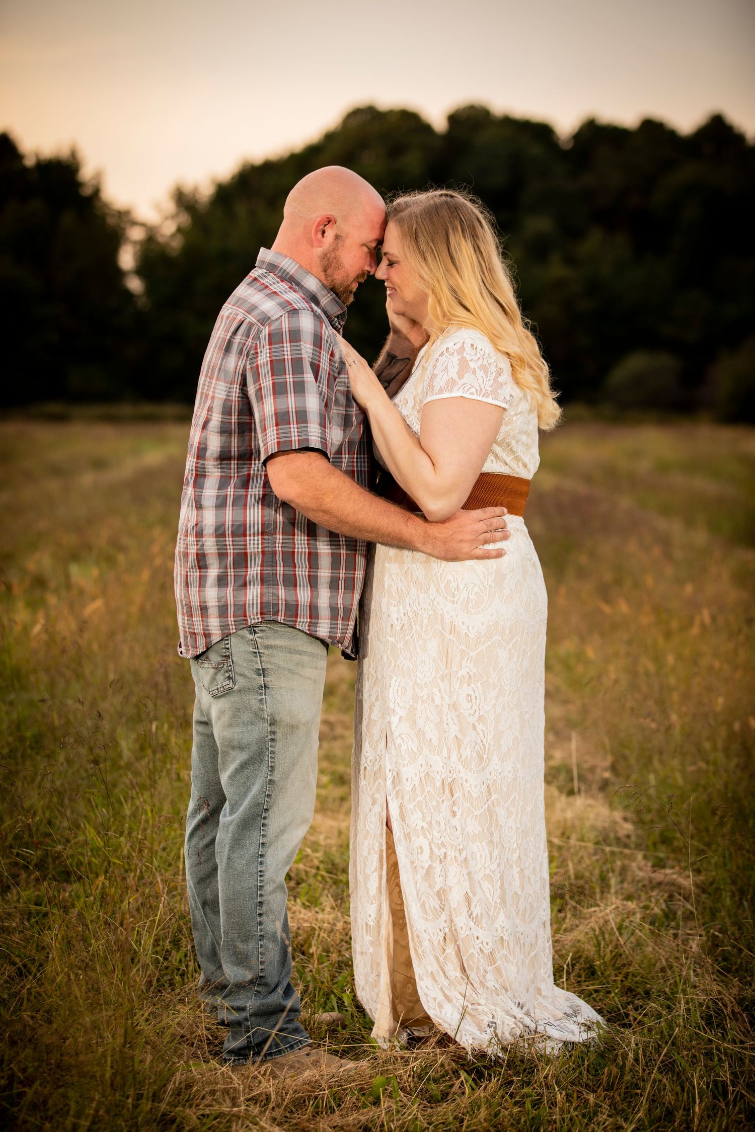 A bald man and blonde woman in white lace dress touch foreheads romantically in a golden field.