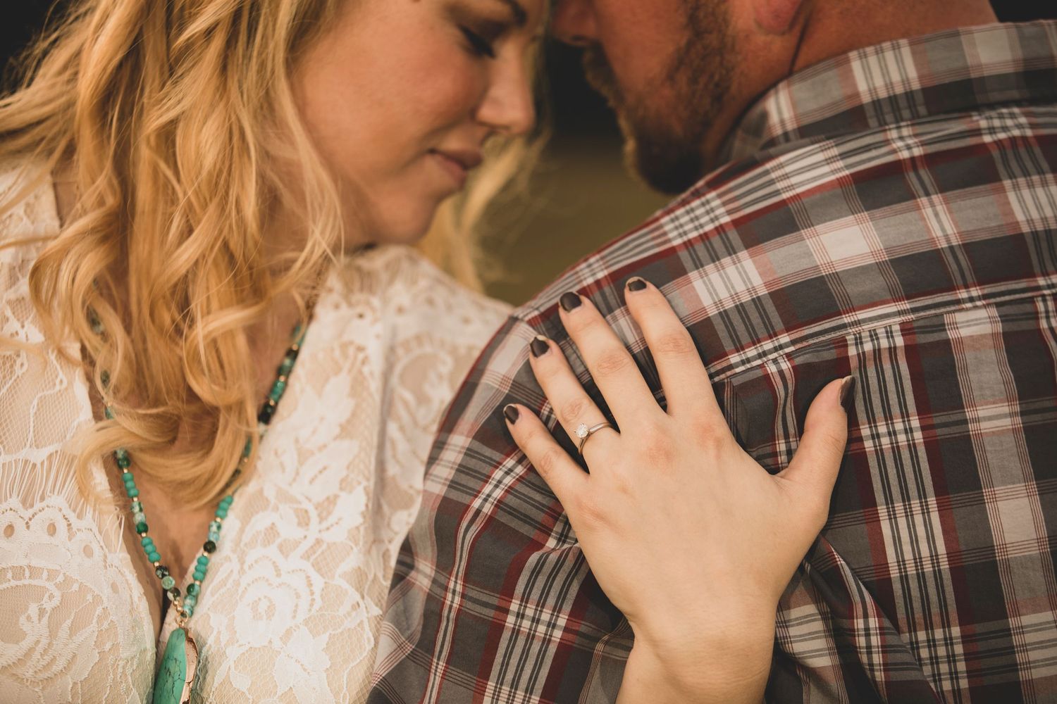 A woman in white lace rests her hand on a mans plaid shirt showcasing an engagement ring.