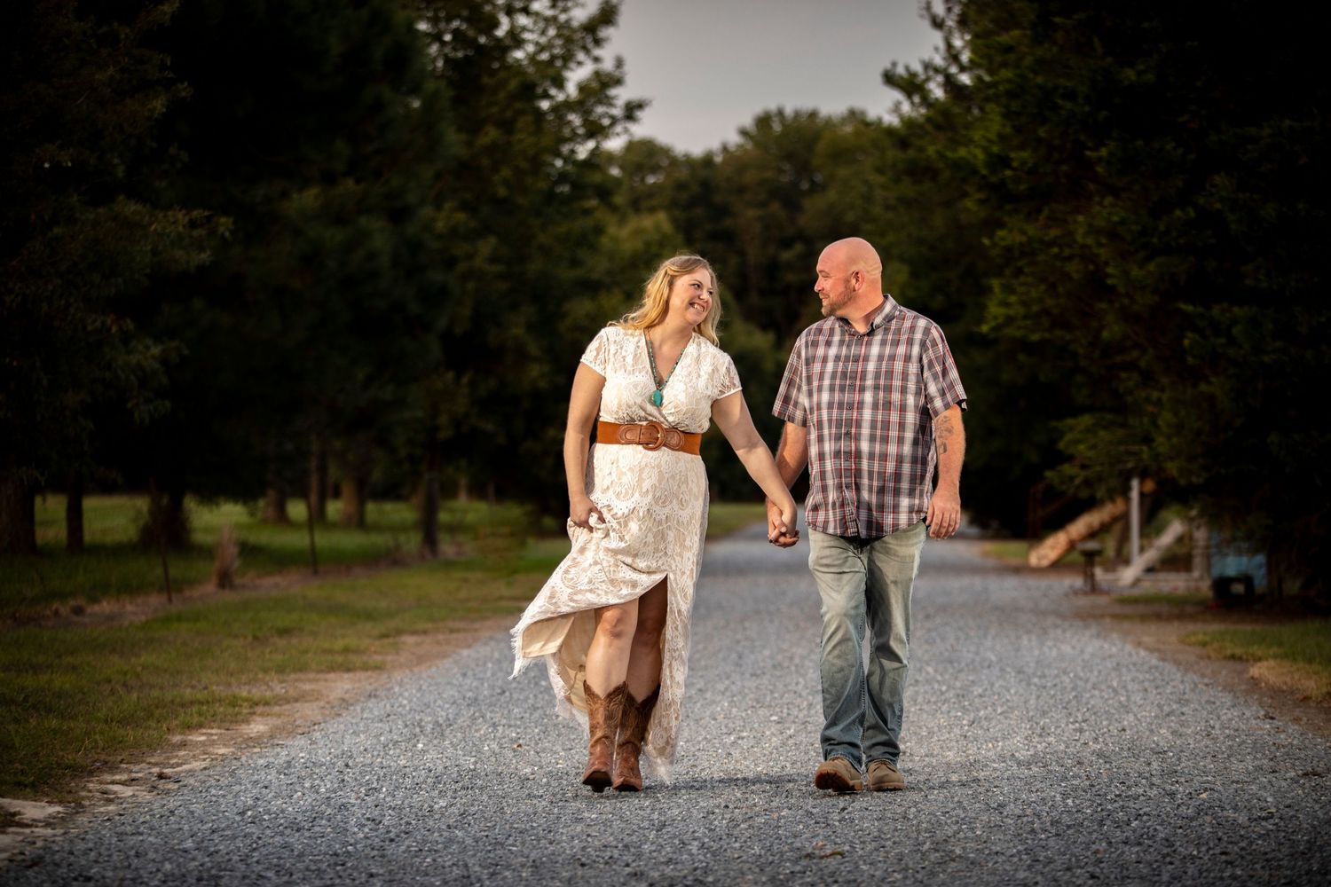 A laughing couple in casual attire holds hands walking down a tree-lined gravel path at sunset.