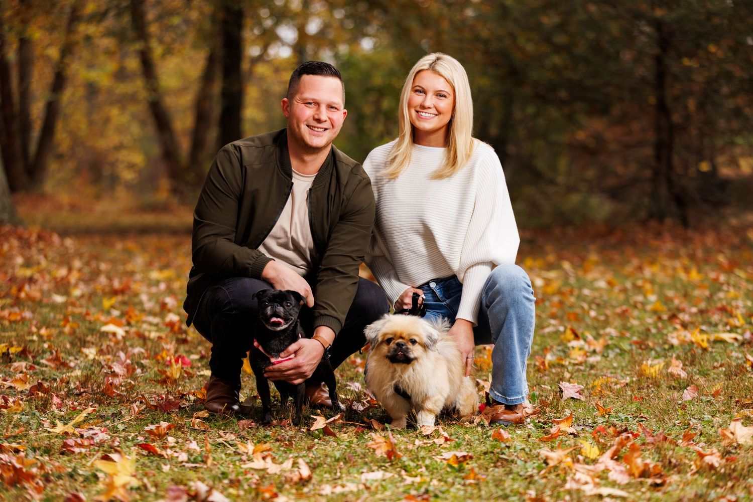Smiling couple crouching with two dogs on colorful autumn leaf-covered ground.