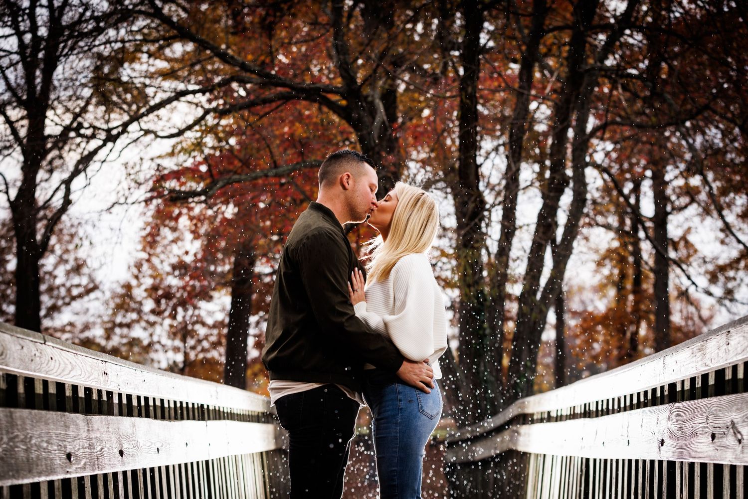 Couple sharing a romantic kiss on a white bridge surrounded by colorful autumn foliage in a park.