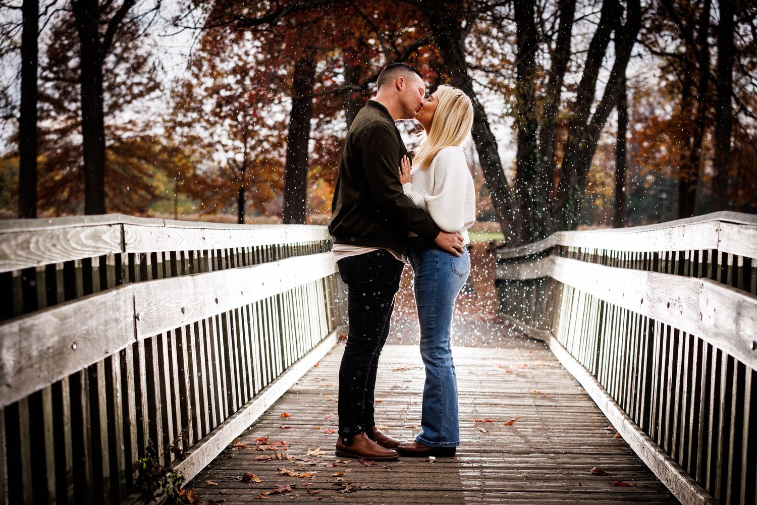 Couple kissing on a wooden bridge with vibrant fall trees and bokeh light effects in background.