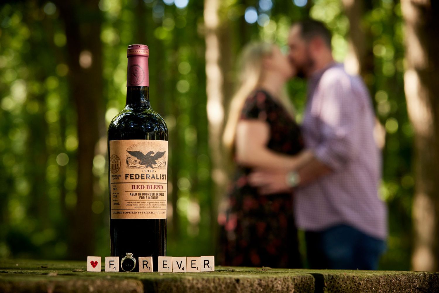 Engagement ring and wine bottle with FOREVER scrabble tiles as couple kisses in blurred forest background.
