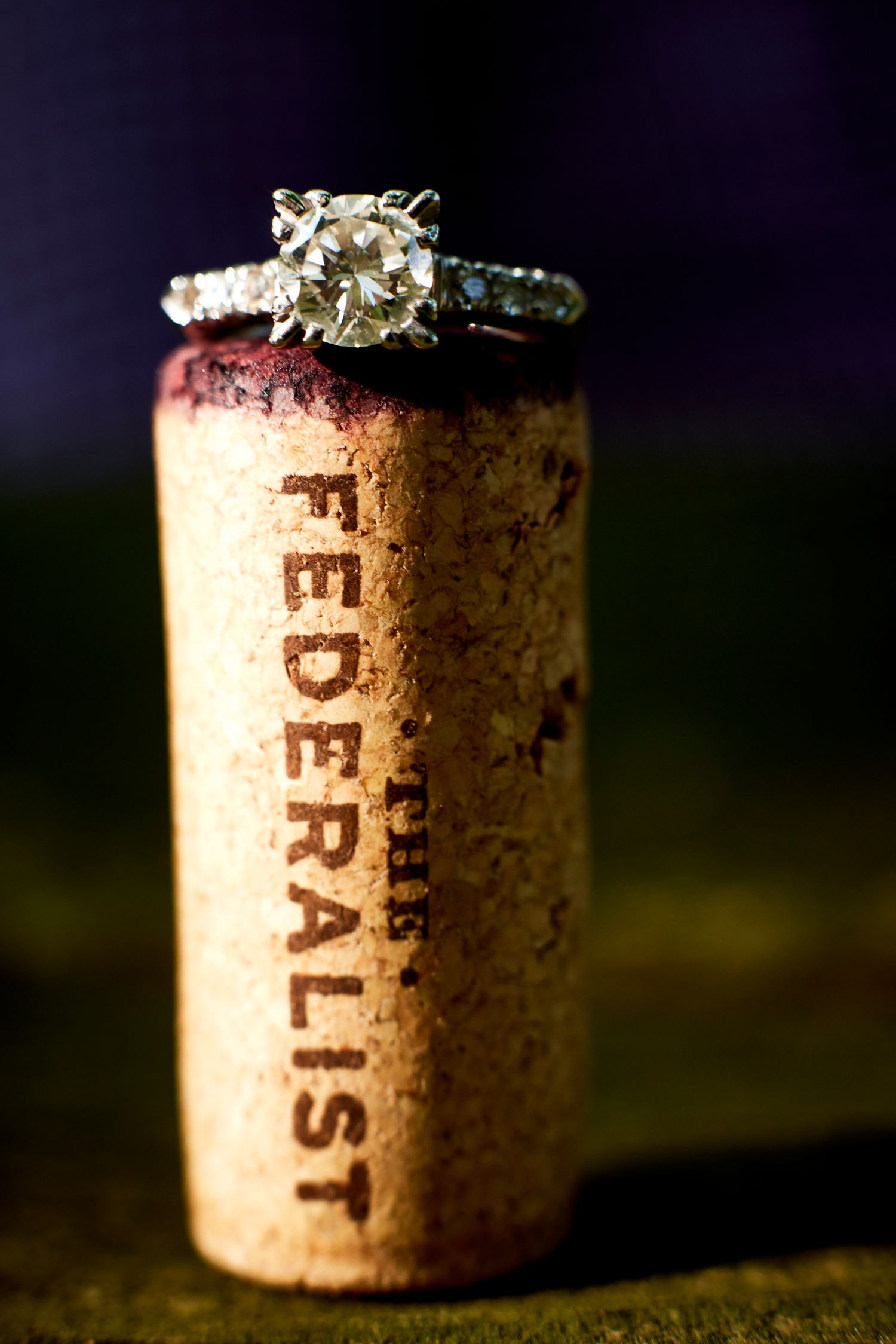 Diamond engagement ring displayed on a Federalist wine cork with dark bokeh background.