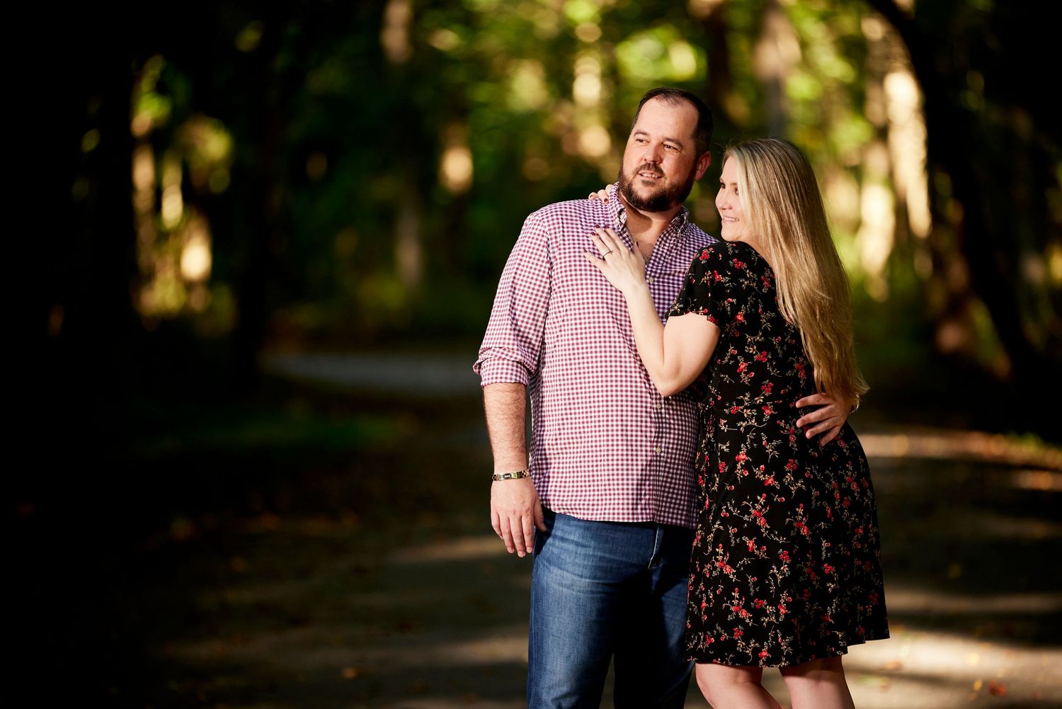 Couple posing together on a tree-lined path, woman in floral dress resting hand on mans chest.