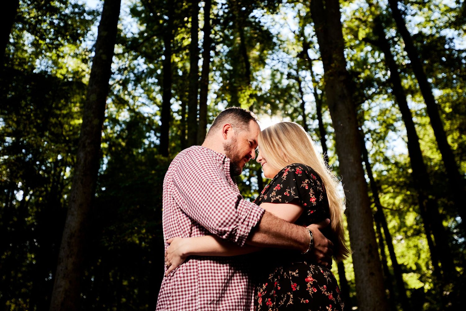 Couple sharing a tender forehead touch embrace surrounded by tall trees in a sunlit forest setting.