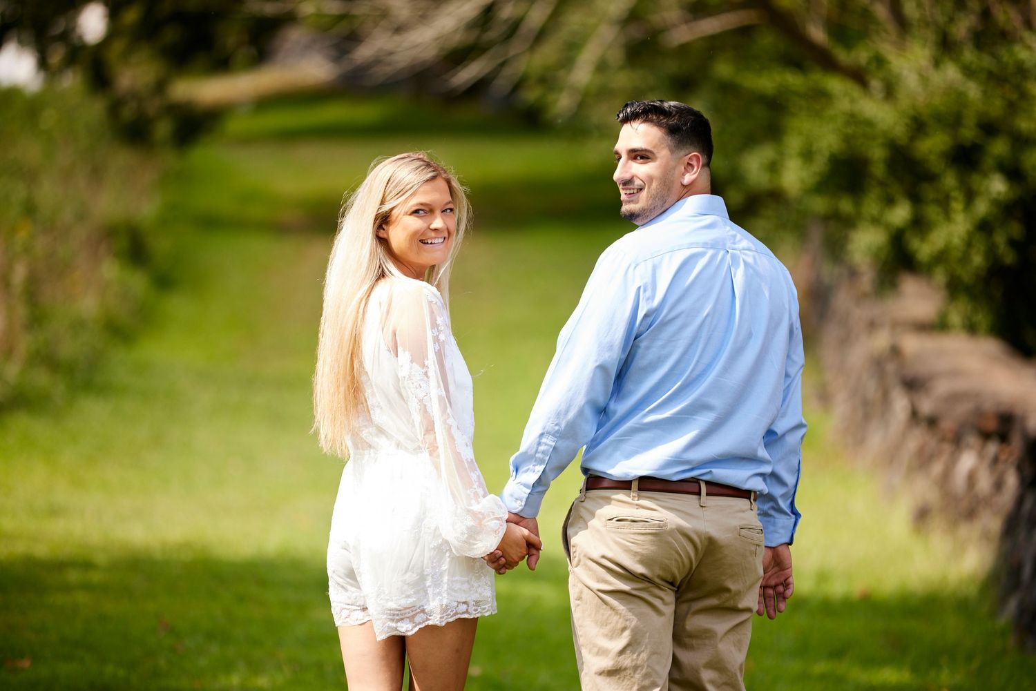 Engaged couple holding hands and looking back while walking along a lush green tree-lined garden path.