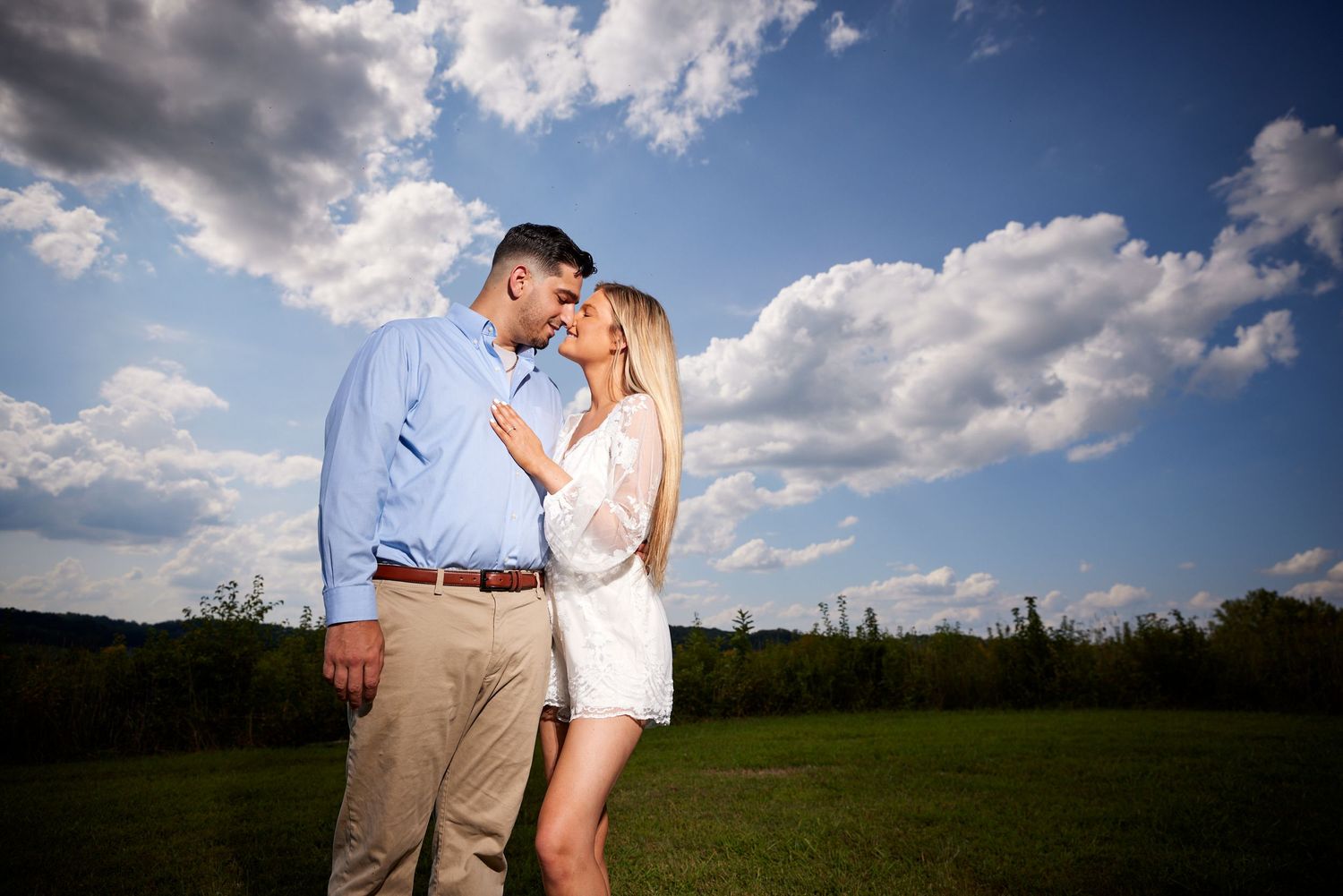 Couple gazing at each other romantically in an open green field under a dramatic blue sky with clouds.